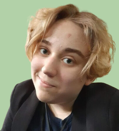 A person with white skin and ear-length blonde hair. They are wearing a purple shirt, and have green eyes and thick eyebrows. They are wearing a black blazer and a dark blue t-shirt.