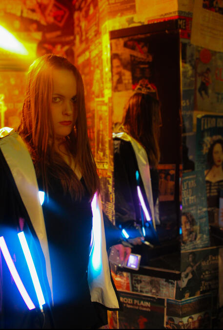 A picture of a woman in a warmly lit room. She has long brown hair, white skin, and is wearing a dark jacket, illuminated by neon lights.