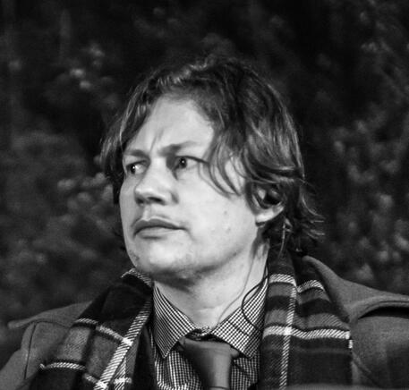 A greyscale picture of a person with whtie skin, chin-length dark hair, a scarf and a suit and tie.