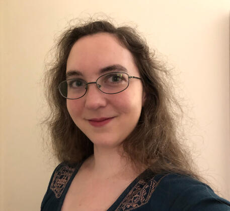 A picture of a woman with white skin, long brown hair, and glasses. She is smiling politely.