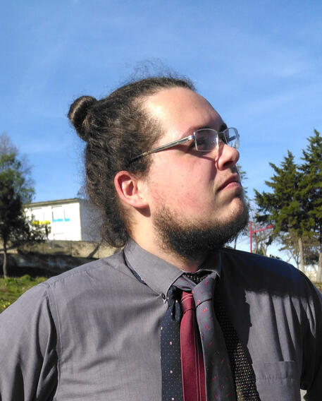 A picture in profile of a man who is standing outside. He has a short brown beard and hair in a bun. He's wearing a formal shirt with three ties and glasses. He is staring introspectively into the distance on a sunny day.